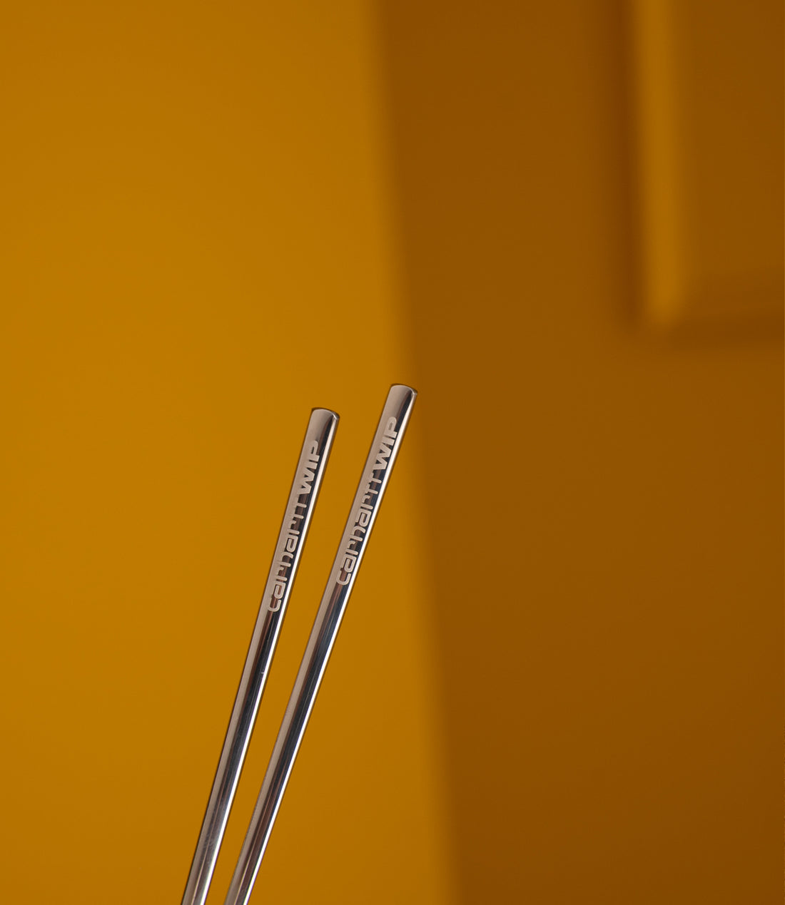 Pair of Carhartt WIP stainless steel chopsticks with engraved logo against an ochre background